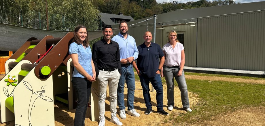 Pfarrerin Kerstin Graf, Sergio Federico vom Fachgebiet 3.1 Bauen, Oberbürgermeister Lennart Siefert, Kitaleiter Christopher Fabeck und Sybil Sasse, Fachgebietsleiterin 2.1 Bildung, Soziales und Sport Fünf Personen stehen nebeneinander auf dem Außengelände der Kita. Links befindet sich ein Spielgerät auf einer Sandfläche, im Hintergrund ist das eingeschossige graue Kita-Gebäude zu sehen. Rund um das Gelände stehen zahlreiche grüne Bäume. Der Himmel ist teilweise bewölkt.