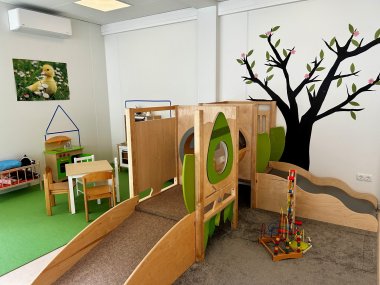 Die Räume am neuen Standort der Kita Kastanienplatz in Friedrichssegen sind kindgerecht eingerichtet und bieten verschiedene Möglichkeiten zum Spielen und Entdecken. Blick in einen hellen Kita-Raum mit einer großen hölzernen Spiel- und Kletterlandschaft mit Rampe, Durchgängen und kleiner Rutsche. An der Wand dahinter ist ein großer schwarzer Baum mit grünen Blättern und rosa Blüten gestaltet. Links stehen eine Kinderküche sowie kleine Tische und Stühle. Auf dem Boden liegen verschiedene Spielmaterialien.