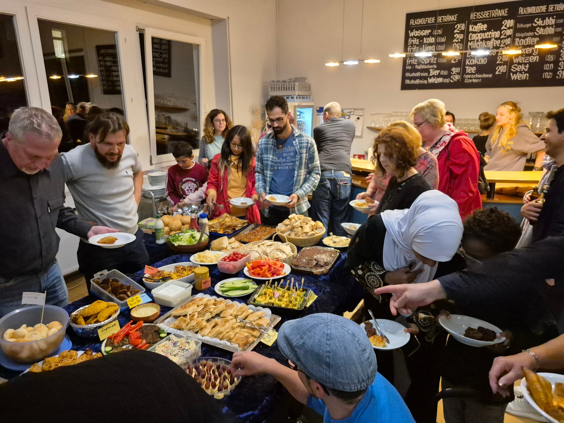 In einem hellen Raum stehen zahlreiche Menschen dicht um ein großes Buffet. Auf dem Tisch sind viele unterschiedliche Speisen angerichtet, darunter Brot und Gebäck, Salate, Gemüse, Dips, kleine Spieße und warme Gerichte. Die Gäste halten Teller in den Händen, bedienen sich am Buffet und unterhalten sich miteinander. Im Hintergrund sind weitere Personen sowie ein Thekenbereich zu sehen.