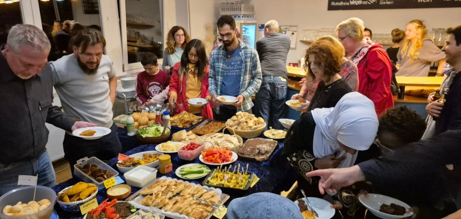 Interkultureller Abend im JUKZ Lahnstein In einem hellen Raum stehen zahlreiche Menschen dicht um ein großes Buffet. Auf dem Tisch sind viele unterschiedliche Speisen angerichtet, darunter Brot und Gebäck, Salate, Gemüse, Dips, kleine Spieße und warme Gerichte. Die Gäste halten Teller in den Händen, bedienen sich am Buffet und unterhalten sich miteinander. Im Hintergrund sind weitere Personen sowie ein Thekenbereich zu sehen.