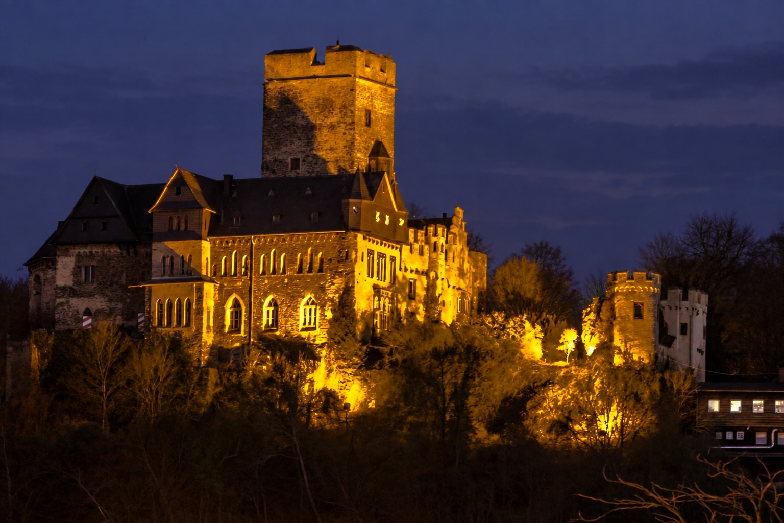 Burg Lahneck bei Einbruch der Dunkelheit. Die Burganlage mit ihrem hohen, quadratischen Bergfried und mehreren Türmen wird von warmem, goldgelbem Licht angestrahlt. Im Vordergrund sind dunkle Bäume und Sträucher zu sehen, im Hintergrund ein dunkelblauer, leicht bewölkter Abendhimmel.