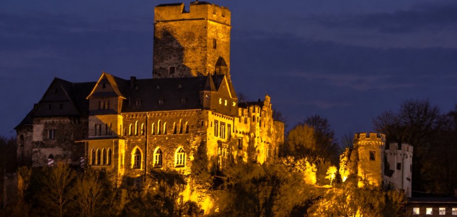 Burg Lahneck Lahnstein Burg Lahneck bei Einbruch der Dunkelheit. Die Burganlage mit ihrem hohen, quadratischen Bergfried und mehreren Türmen wird von warmem, goldgelbem Licht angestrahlt. Im Vordergrund sind dunkle Bäume und Sträucher zu sehen, im Hintergrund ein dunkelblauer, leicht bewölkter Abendhimmel.