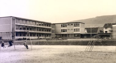 Historische Schwarz-Weiß-Außenaufnahme eines mehrteiligen, dreigeschossigen Schulgebäudes mit zahlreichen Fenstern. Im Vordergrund befindet sich ein großer Schulhof mit mehreren Schaukeln und einem gebogenen Klettergerüst. Drei Kinder befinden sich bei den Schaukeln. Hinter dem Gebäude ist ein bewaldeter Hang zu sehen.