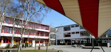 Farbaufnahme eines Schulhofs mit einem mehrteiligen, dreigeschossigen Schulgebäude. Die weißen Fassaden haben zahlreiche rot gerahmte Fenster. Vor dem Gebäude stehen mehrere Bäume und Sträucher, auf dem gepflasterten Schulhof liegen große Steine. Rechts überspannt ein großes rot-weiß gestreiftes Sonnensegel einen Teil des Hofes.
