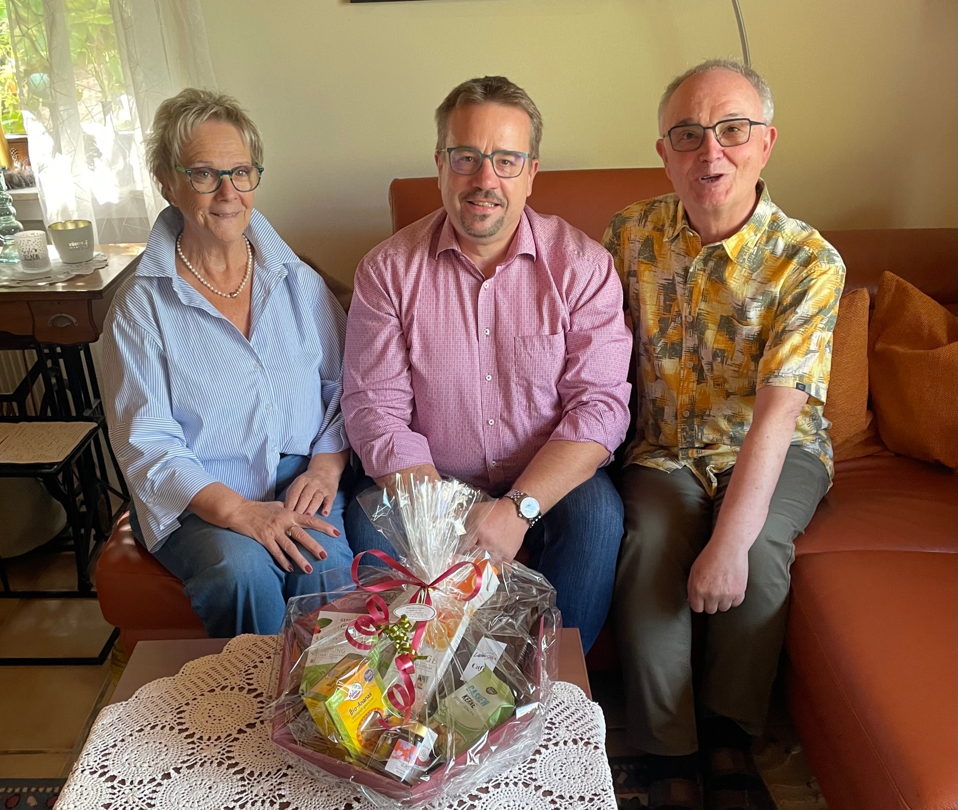 Drei Personen sitzen nebeneinander auf einem orangefarbenen Sofa in einem Wohnzimmer und blicken in die Kamera. Links sitzt eine Frau mit kurzem blond-grauem Haar, Brille, hellblau gestreifter Bluse und Perlenkette. In der Mitte sitzt ein Mann mit Brille, kurzem dunklem Haar und Bart, der ein rot-weiß kariertes Hemd trägt. Rechts sitzt ein Mann mit Brille, kurzem grauem Haar und einem gelb-grau gemusterten Kurzarmhemd. Vor den drei Personen steht auf einem Tisch ein in Folie verpackter Geschenkkorb mit roter Schleife.