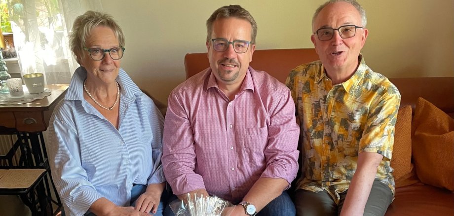Ehejubiläum Mallmann Drei Personen sitzen nebeneinander auf einem orangefarbenen Sofa in einem Wohnzimmer und blicken in die Kamera. Links sitzt eine Frau mit kurzem blond-grauem Haar, Brille, hellblau gestreifter Bluse und Perlenkette. In der Mitte sitzt ein Mann mit Brille, kurzem dunklem Haar und Bart, der ein rot-weiß kariertes Hemd trägt. Rechts sitzt ein Mann mit Brille, kurzem grauem Haar und einem gelb-grau gemusterten Kurzarmhemd. Vor den drei Personen steht auf einem Tisch ein in Folie verpackter Geschenkkorb mit roter Schleife.