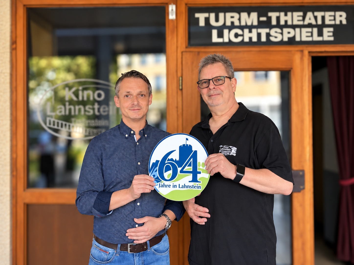 Zwei Männer stehen vor dem Eingang des Kino Lahnstein. Über der Eingangstür ist ein schwarzes Schild mit der weißen Aufschrift „Turm-Theater Lichtspiele“ zu sehen, auf einer Glasscheibe links daneben der Schriftzug „Kino Lahnstein“. Gemeinsam halten die beiden Männer ein rundes Schild in den Händen, auf dem groß die Zahl 64 und darunter „Jahre in Lahnstein“ steht.