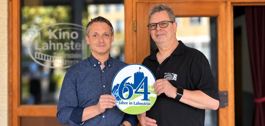 Lahnstein-Label Zwei Männer stehen vor dem Eingang des Kino Lahnstein. Über der Eingangstür ist ein schwarzes Schild mit der weißen Aufschrift „Turm-Theater Lichtspiele“ zu sehen, auf einer Glasscheibe links daneben der Schriftzug „Kino Lahnstein“. Gemeinsam halten die beiden Männer ein rundes Schild in den Händen, auf dem groß die Zahl 64 und darunter „Jahre in Lahnstein“ steht.