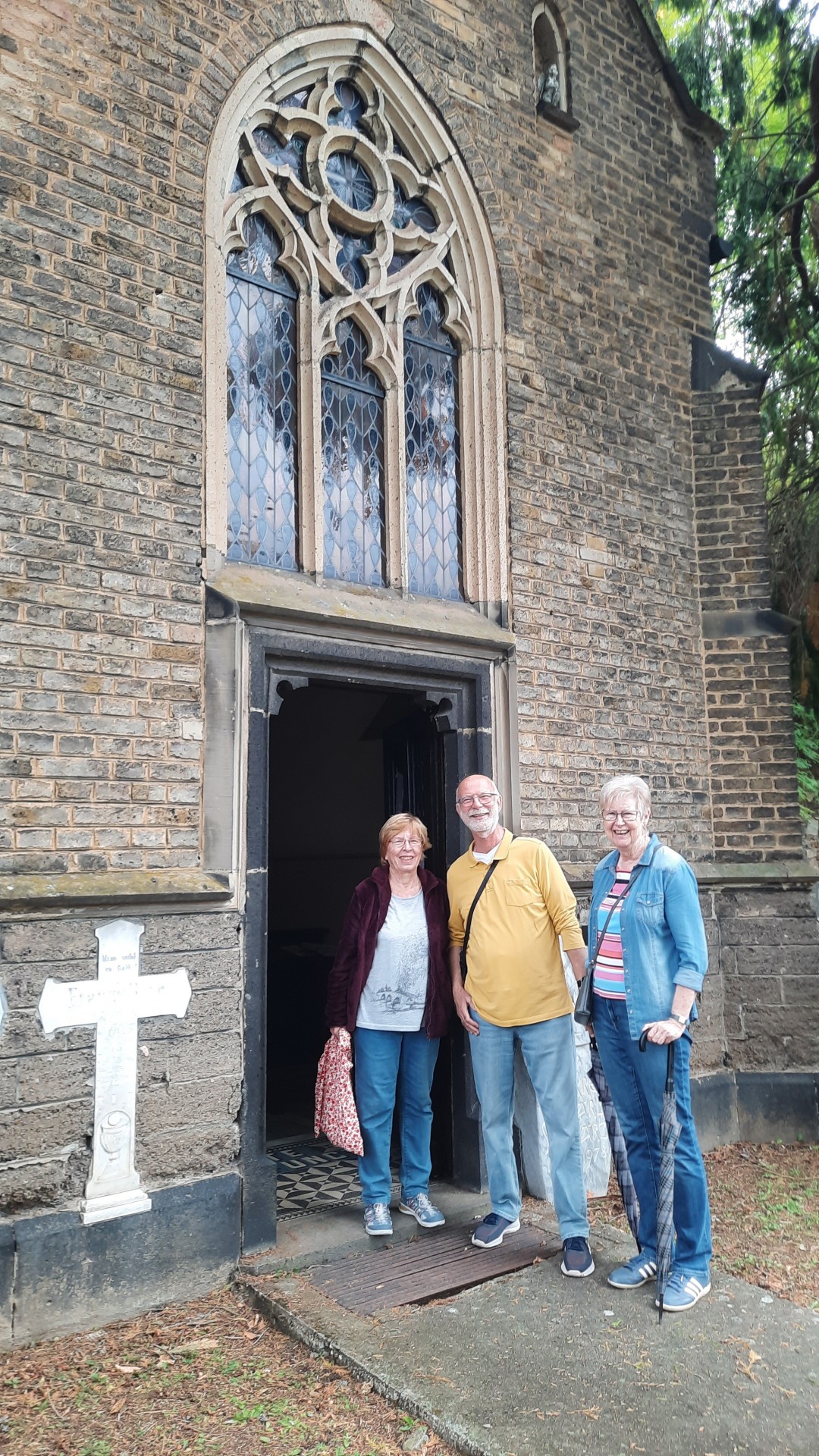 Drei Personen stehen vor dem geöffneten Eingang einer historischen Gruftkapelle aus dunklem Backstein. Über der Eingangstür befindet sich ein großes spitzbogiges Fenster mit steinernem Maßwerk und verglasten Feldern. Links neben dem Eingang steht ein weißes steinernes Kreuz. Die Kapelle ist von Bäumen und Grün umgeben.