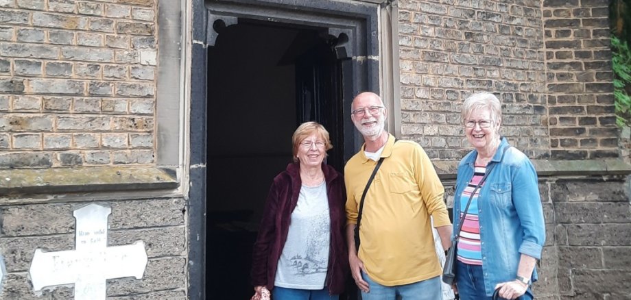 Tag des offenen Denkmals Drei Personen stehen vor dem geöffneten Eingang einer historischen Gruftkapelle aus dunklem Backstein. Über der Eingangstür befindet sich ein großes spitzbogiges Fenster mit steinernem Maßwerk und verglasten Feldern. Links neben dem Eingang steht ein weißes steinernes Kreuz. Die Kapelle ist von Bäumen und Grün umgeben.