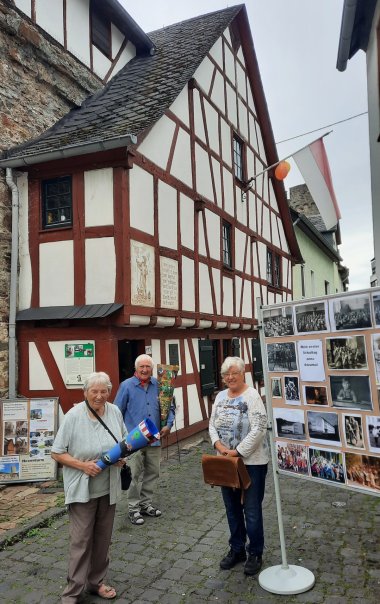 Tag des offenen Denkmals Vor einem historischen Fachwerkhaus mit rotbraunen Balken und weißen Gefachen stehen drei Personen auf einer gepflasterten Gasse. Eine Frau hält eine bunt gestaltete Schultüte, der Mann in der Mitte eine weitere Schultüte. Rechts neben der Gruppe steht eine Ausstellungstafel mit historischen Schwarz-Weiß-Fotografien. Vor und neben dem Gebäude befinden sich weitere Informationstafeln. Am Fachwerkhaus hängt eine rot-weiße Fahne.
