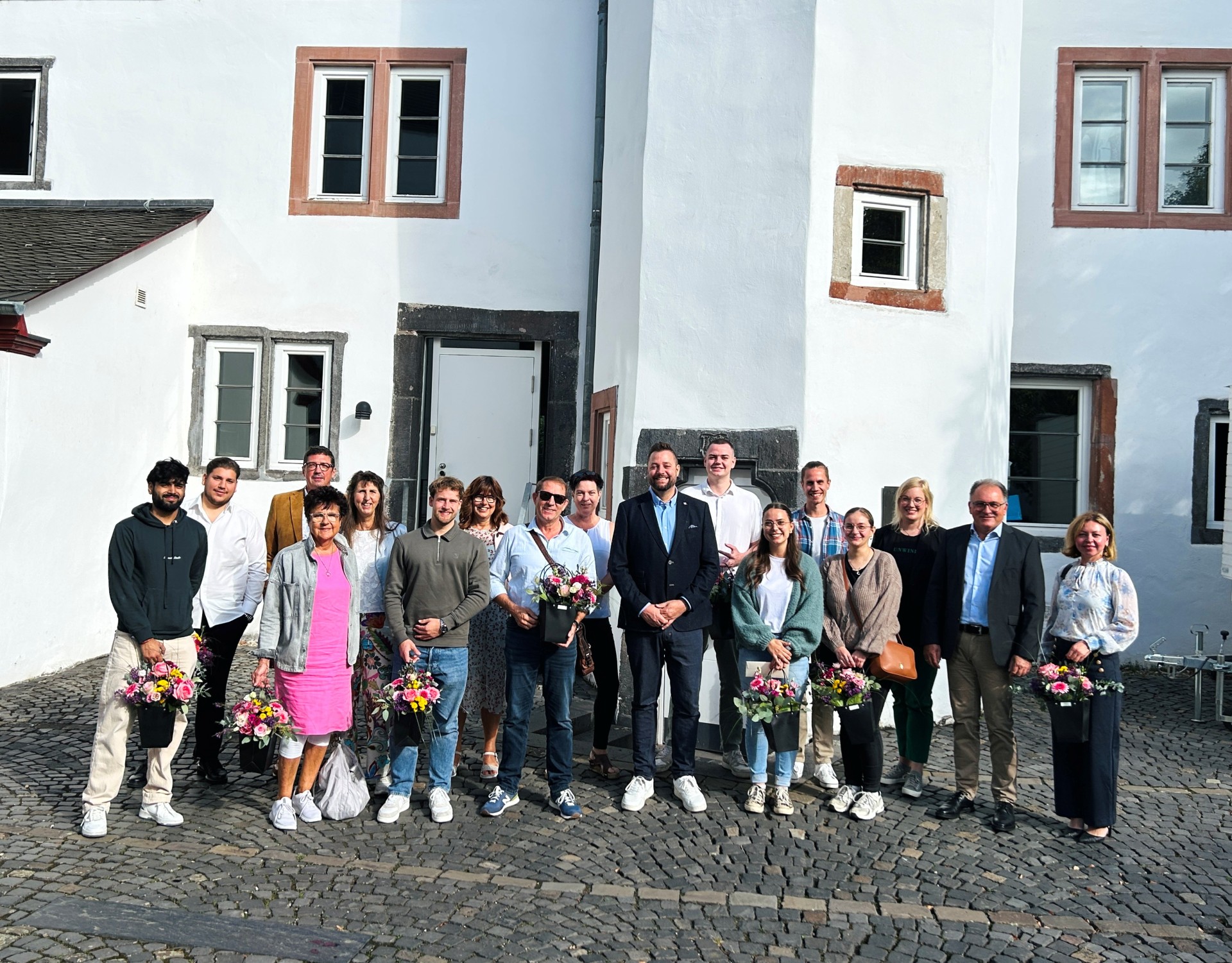 Eine größere Gruppe von Frauen und Männern steht auf einem gepflasterten Platz vor einem weißen historischen Gebäude. Mehrere Personen halten Blumensträuße in den Händen. Die Gruppe steht in zwei Reihen nebeneinander und blickt in die Kamera.