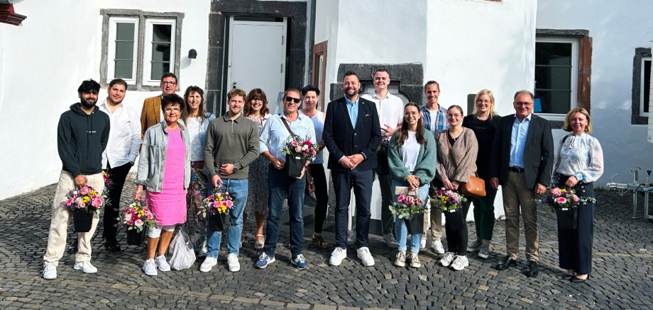 Eine größere Gruppe von Frauen und Männern steht auf einem gepflasterten Platz vor einem weißen historischen Gebäude. Mehrere Personen halten Blumensträuße in den Händen. Die Gruppe steht in zwei Reihen nebeneinander und blickt in die Kamera.