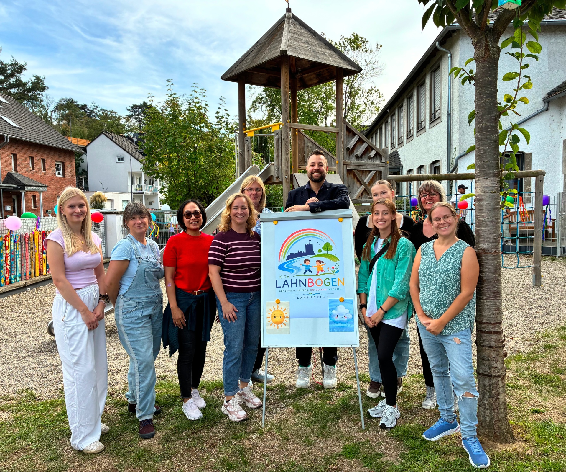 Eine Gruppe von zehn Frauen und einem Mann steht auf dem Außengelände der Kita Lahnbogen vor einem Schild mit dem neuen Namen und Logo der Einrichtung. Im Hintergrund befinden sich ein hölzernes Klettergerüst mit Rutsche, weitere Spielgeräte und das Kita-Gebäude.