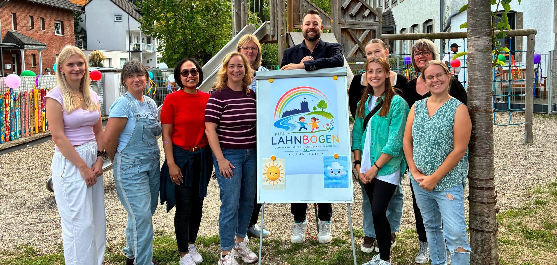 Eine Gruppe von zehn Frauen und einem Mann steht auf dem Außengelände der Kita Lahnbogen vor einem Schild mit dem neuen Namen und Logo der Einrichtung. Im Hintergrund befinden sich ein hölzernes Klettergerüst mit Rutsche, weitere Spielgeräte und das Kita-Gebäude.