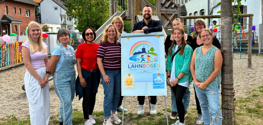Kita lahnbogen Lahnstein Eine Gruppe von zehn Frauen und einem Mann steht auf dem Außengelände der Kita Lahnbogen vor einem Schild mit dem neuen Namen und Logo der Einrichtung. Im Hintergrund befinden sich ein hölzernes Klettergerüst mit Rutsche, weitere Spielgeräte und das Kita-Gebäude.