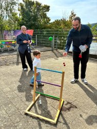 Kita Lahnbogen Lahnstein Ein Mann und eine Frau stehen gemeinsam mit einem kleinen Jungen auf dem Außengelände der Kita. Der Mann hält ein Wurfspiel mit roten Kugeln in der Hand, vor ihnen steht ein Gestell mit drei farbigen Querstreben. Im Hintergrund sind Bäume und weitere Spielangebote zu sehen.