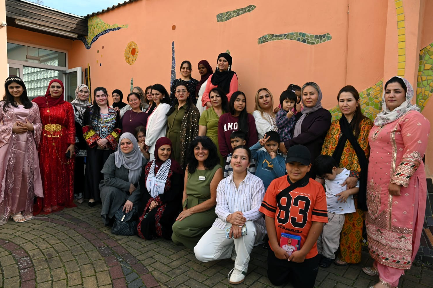 Gruppenfoto von Frauen und mehreren Kindern vor einer orangefarbenen Hauswand mit bunten Mosaikelementen. Einige Frauen tragen traditionelle Kleidung oder Kopftücher, andere Alltagskleidung. Ein Teil der Gruppe steht, weitere Personen sitzen oder knien in der ersten Reihe.