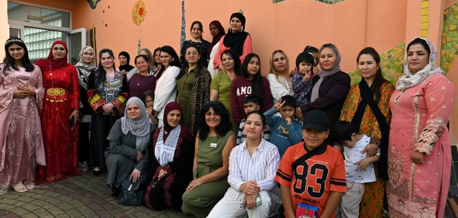 Zehn Jahre Interkulturelles Frauencafé Lahnstein Gruppenfoto von Frauen und mehreren Kindern vor einer orangefarbenen Hauswand mit bunten Mosaikelementen. Einige Frauen tragen traditionelle Kleidung oder Kopftücher, andere Alltagskleidung. Ein Teil der Gruppe steht, weitere Personen sitzen oder knien in der ersten Reihe.
