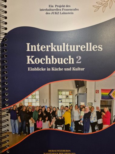 Frisch erschienen: Das zweite interkulturelle Kochbuch des Frauencafés vereint Rezepte und persönliche Geschichten der beteiligten Frauen. Nahaufnahme des spiralgebundenen „Interkulturellen Kochbuchs 2 – Einblicke in Küche und Kultur“. Auf dem dunkelblauen Cover ist ein Gruppenfoto von Frauen und Kindern im Jugendkulturzentrum Lahnstein zu sehen.