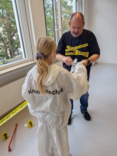 Beim Workshop zeigte Privatdetektiv Alexander Schrumpf den Kindern vieles rund um den Detektivberuf. Ein Mädchen steht mit dem Rücken zur Kamera und trägt einen weißen Schutzanzug mit der Aufschrift „Kriminaltechnik“. Vor ihr steht Detektiv Alexander Schrumpf in einem schwarzen T-Shirt. Gemeinsam arbeiten sie an einem Gegenstand. Auf dem Boden daneben ist ein mit gelbem Absperrband markierter Bereich mit nummerierten Hinweisschildern und verschiedenen Gegenständen zu sehen.