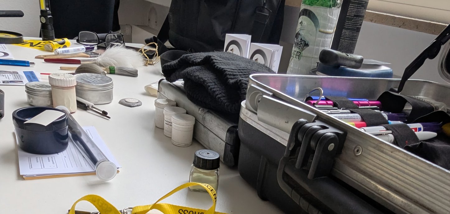 Auf einem weißen Tisch liegen zahlreiche Detektiv- und Ermittlungsutensilien. Im Vordergrund befinden sich geöffnete Handschellen, daneben ein gelbes Band mit der Aufschrift „Crime Scene – Do Not Cross“ sowie verschiedene kleine Behälter, Mess- und Arbeitsgeräte. Rechts steht ein geöffneter Koffer mit weiteren Materialien.