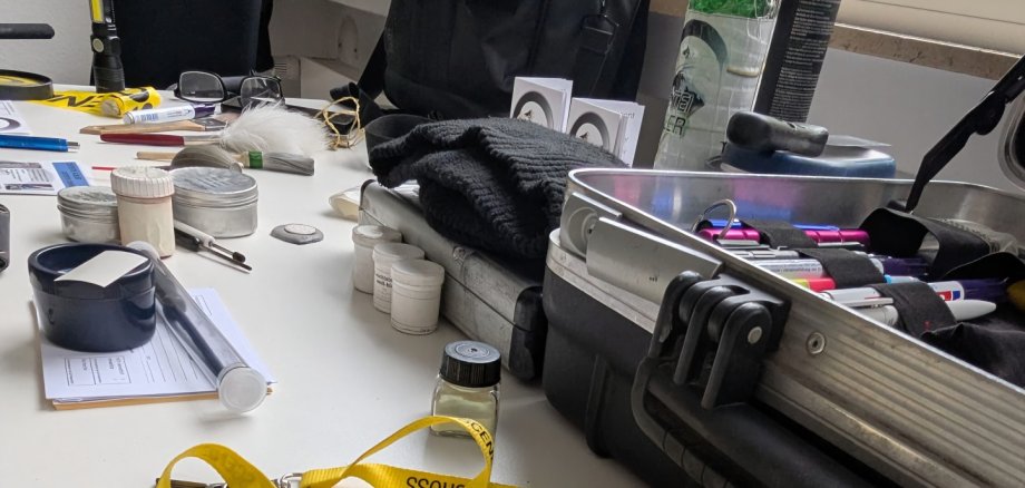 Verschiedene Utensilien aus dem Arbeitsalltag eines Detektivs Auf einem weißen Tisch liegen zahlreiche Detektiv- und Ermittlungsutensilien. Im Vordergrund befinden sich geöffnete Handschellen, daneben ein gelbes Band mit der Aufschrift „Crime Scene – Do Not Cross“ sowie verschiedene kleine Behälter, Mess- und Arbeitsgeräte. Rechts steht ein geöffneter Koffer mit weiteren Materialien.