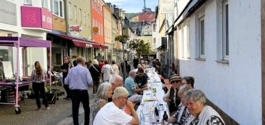 Eine lange Tafel mit Bierzeltgarnituren steht in einer gepflasterten Straße zwischen Häusern und Geschäften. Zahlreiche Menschen sitzen gemeinsam an den Tischen, weitere Personen stehen oder gehen durch die Straße.