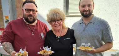 Drei Personen stehen nebeneinander auf einer gepflasterten Fläche und halten Teller mit verschiedenen nummerierten Zwiebelkuchenstücken in den Händen. Im Hintergrund sind orangefarbene Bänke und weitere Teller auf einem Tisch zu sehen.