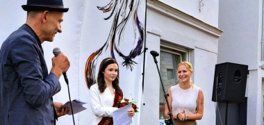 Ein Mann mit Hut und Mikrofon steht auf einer kleinen Bühne neben zwei Frauen. Eine der Frauen trägt ein langes weißes Kleid und eine rote Schärpe und hält einen Blumenstrauß sowie ein Blatt Papier in den Händen. Im Hintergrund sind eine Gitarre, Mikrofonständer und Lautsprecher zu sehen.