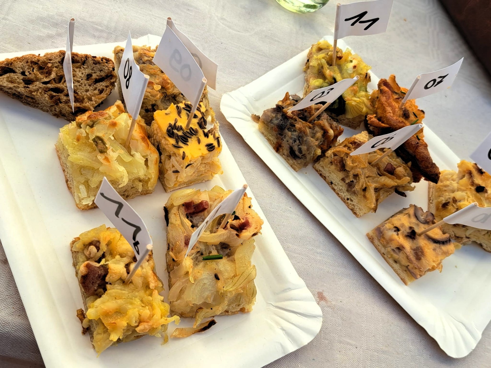 Auf zwei weißen Papptellern liegen verschiedene kleine Stücke Zwiebelkuchen. Die einzelnen Kostproben sind mit kleinen nummerierten Papierfähnchen gekennzeichnet.
