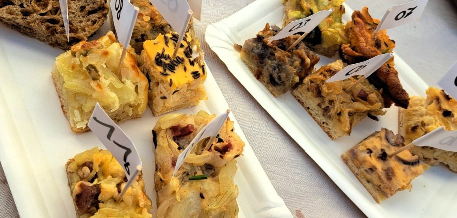 Auf zwei weißen Papptellern liegen verschiedene kleine Stücke Zwiebelkuchen. Die einzelnen Kostproben sind mit kleinen nummerierten Papierfähnchen gekennzeichnet.