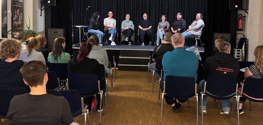 Raum für Austausch Blick in einen Veranstaltungsraum des Jugendkulturzentrums Lahnstein. Im Vordergrund sitzt das Publikum auf Stühlen, auf der Bühne sitzen sieben Personen nebeneinander und sprechen miteinander.