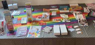 Infomaterial Queer Ein Tisch mit zahlreichen Informationsmaterialien rund um queere Themen. Auf einer Regenbogen- und einer Progress-Pride-Flagge liegen Broschüren, Flyer, Postkarten, Aufkleber, Stifte und weitere Materialien zu sexueller und geschlechtlicher Vielfalt.