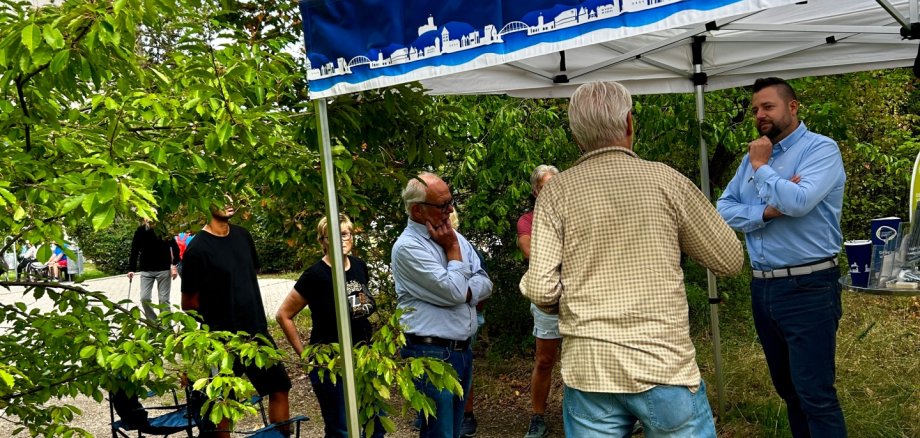 Lennart Live im Kurpark Mehrere Personen stehen im Kurpark unter und neben einem blau-weißen Pavillon und unterhalten sich miteinander. Rund um die Gruppe wachsen zahlreiche Bäume und Sträucher. Im Vordergrund sind Äste und Grünflächen zu sehen.