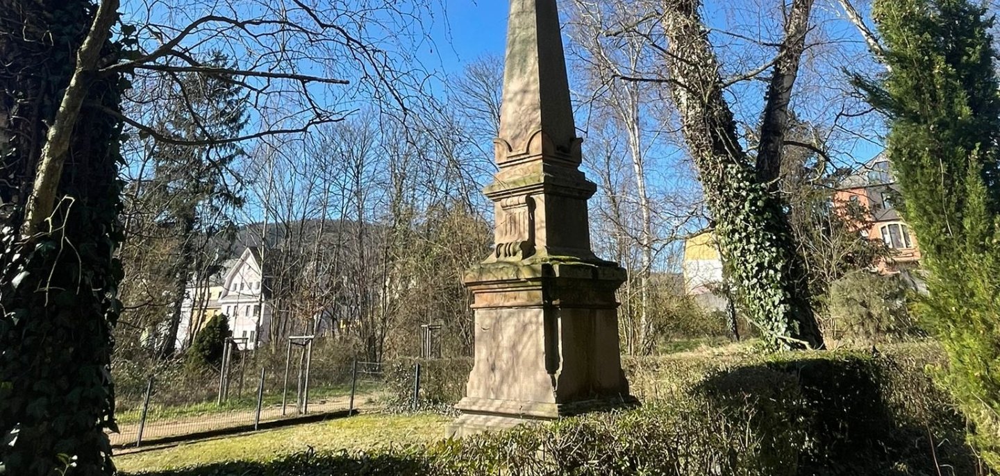 Ein historisches steinernes Denkmal mit hohem Obelisken steht in einer kleinen Parkanlage, umgeben von Hecken und hohen, teilweise kahlen Bäumen. Die Aufnahme entstand bei strahlendem Sonnenschein unter einem klaren blauen Himmel. Im Hintergrund sind einzelne Wohnhäuser zwischen den Bäumen zu erkennen. Die Perspektive von unten lässt das Denkmal besonders imposant wirken.