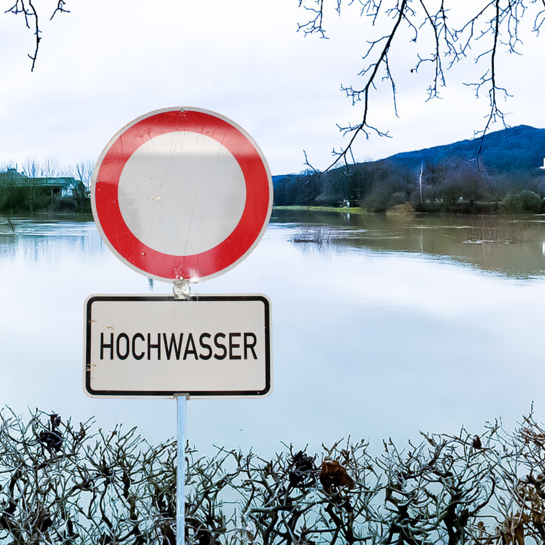 Ein "Durchfahrt-verboten-Schild" mit dem Zusatzwarnhinweis "Hochwasser", das unmittelbar vor einem überschwemmten Bereich steht.