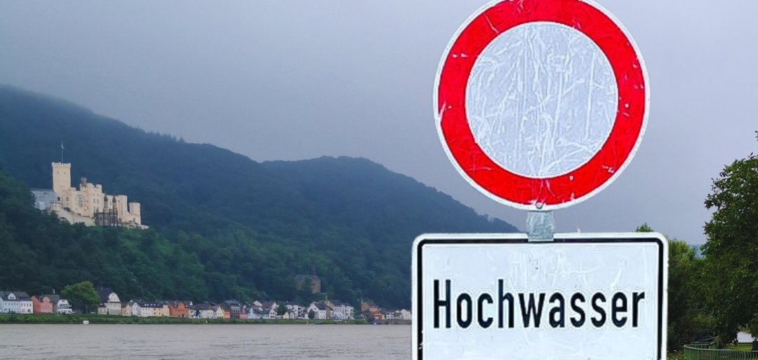 Ein "Durchfahrt-verboten-Schild" mit dem Zusatzwarnhinweis "Hochwasser", das unmittelbar vor einem überschwemmten Bereich des Rheins steht. Im Hintergrund ist Schloss Stolzenfels zu sehen.
