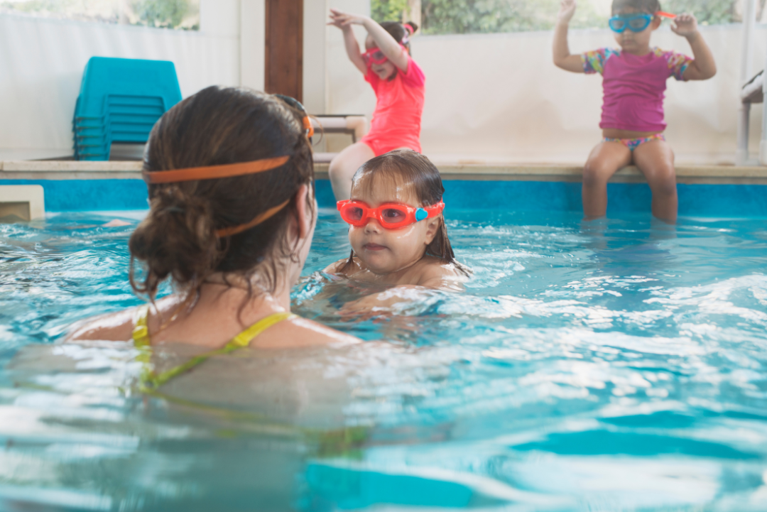 Eine erwachsene Frau von hinten vor einem Mädchen mit Taucherbrille in einem Schwimmbecken. Im Hintergrund sitzen zwei weitere Kinder am Beckenrand.