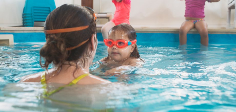 Eine erwachsene Frau von hinten vor einem Mädchen mit Taucherbrille in einem Schwimmbecken. Im Hintergrund sitzen zwei weitere Kinder am Beckenrand.