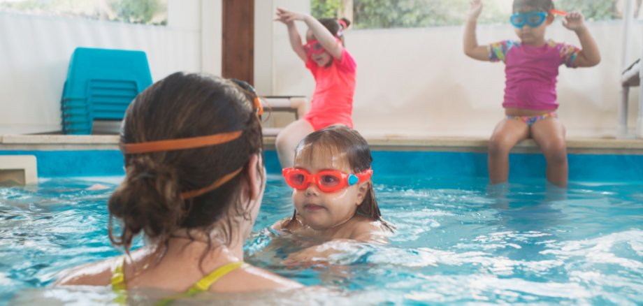 Eine erwachsene Frau von hinten vor einem Mädchen mit Taucherbrille in einem Schwimmbecken. Im Hintergrund sitzen zwei weitere Kinder am Beckenrand.