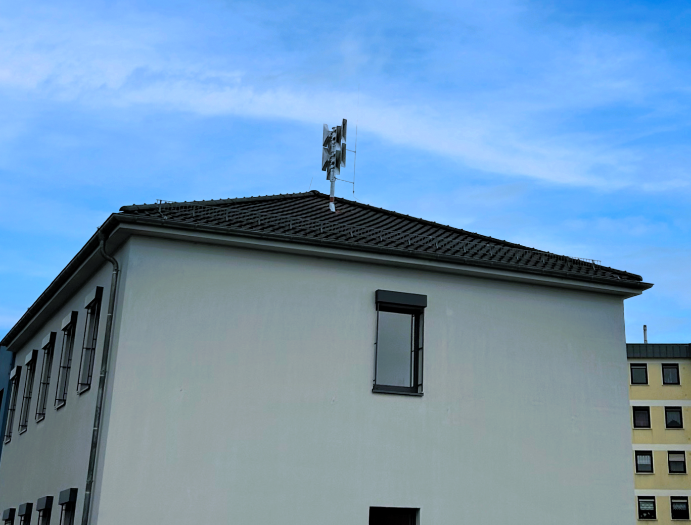 Auf dem Dach eines hellen Gebäudes ist eine moderne Sirene montiert. Sie ragt vor einem wolkenlosen blauen Himmel empor. Man erkennt einen Teil des weißen Gebäudes mit Fenster, im Hintergrund ist ein gelbes Haus zu sehen.