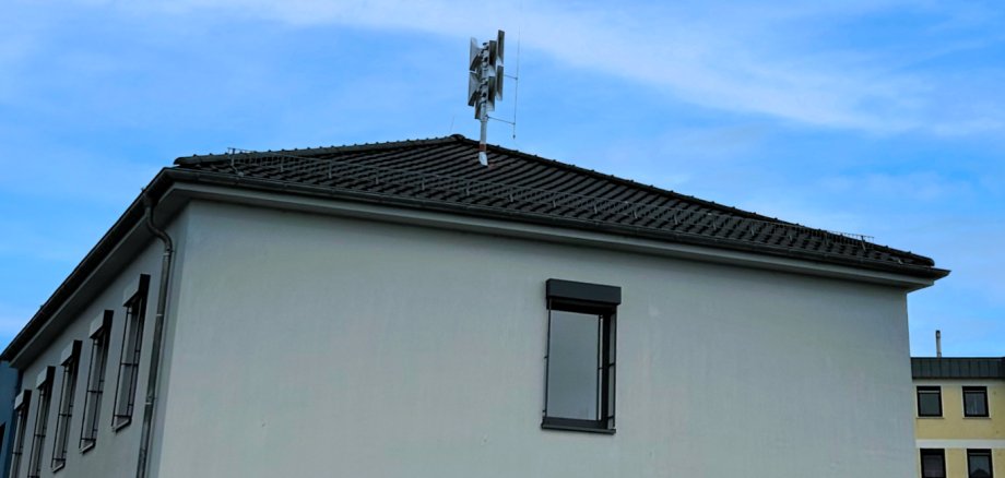 Auf dem Dach eines hellen Gebäudes ist eine moderne Sirene montiert. Sie ragt vor einem wolkenlosen blauen Himmel empor. Man erkennt einen Teil des weißen Gebäudes mit Fenster, im Hintergrund ist ein gelbes Haus zu sehen.