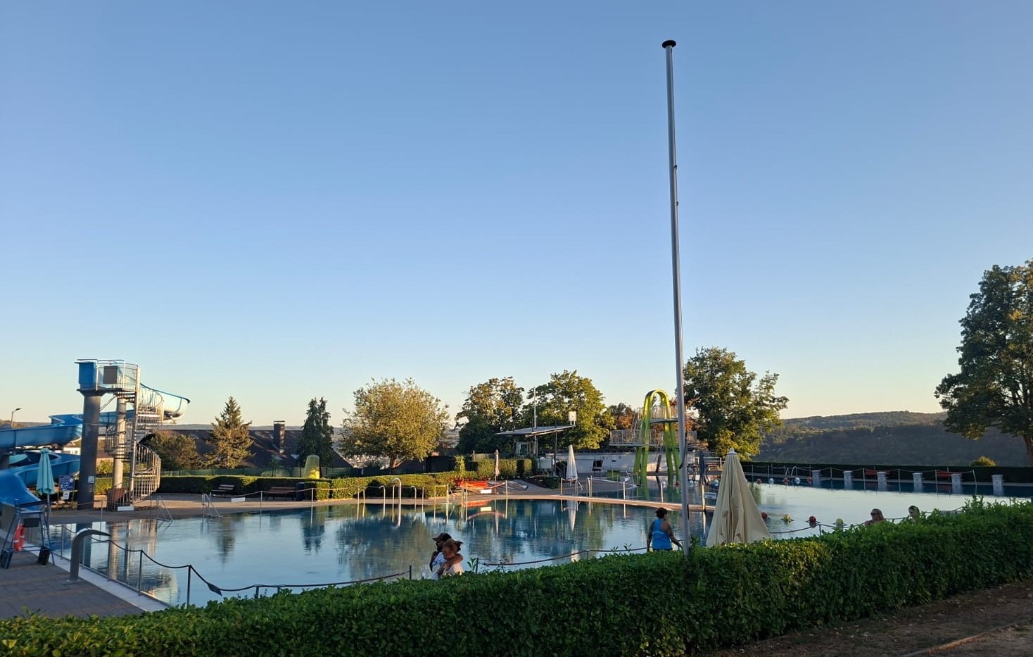 Blick über das Lahnsteiner Freibad bei sonnigem Wetter und blauem Himmel. Im Mittelpunkt liegt das große Schwimmbecken, in dem sich einige Badegäste aufhalten. Links ist die blaue Wasserrutsche mit ihrem Treppenturm zu sehen, im hinteren Bereich eine weitere Rutschenanlage. Rund um die Becken befinden sich Sonnenschirme, Sitz- und Liegebereiche sowie grüne Hecken und Bäume. Im Hintergrund erstreckt sich die bewaldete Landschaft rund um Lahnstein.