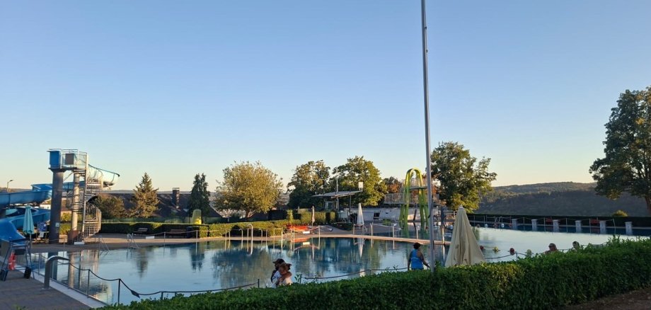 Freibad Lahnstein Blick über das Lahnsteiner Freibad bei sonnigem Wetter und blauem Himmel. Im Mittelpunkt liegt das große Schwimmbecken, in dem sich einige Badegäste aufhalten. Links ist die blaue Wasserrutsche mit ihrem Treppenturm zu sehen, im hinteren Bereich eine weitere Rutschenanlage. Rund um die Becken befinden sich Sonnenschirme, Sitz- und Liegebereiche sowie grüne Hecken und Bäume. Im Hintergrund erstreckt sich die bewaldete Landschaft rund um Lahnstein.