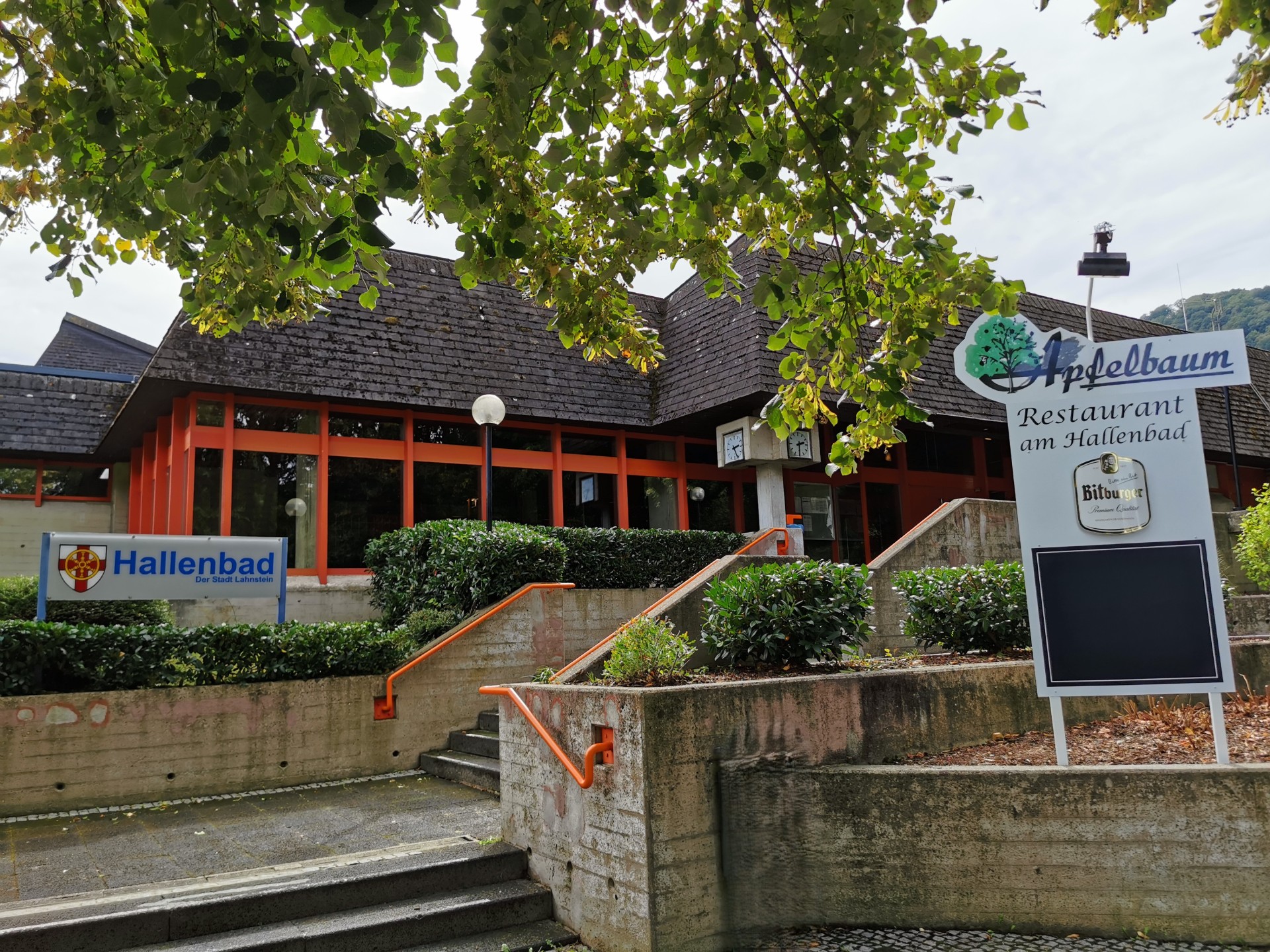 Außenansicht eines flachen Gebäudes mit dunklem Schrägdach und einer Fassade mit großen Fensterflächen und orangefarbenen Rahmen. Eine breite Treppenanlage mit orangefarbenen Handläufen führt zum Eingang hinauf. Vor dem Gebäude stehen Sträucher und Bäume. Links befindet sich ein Schild mit der Aufschrift „Hallenbad der Stadt Lahnstein“, rechts eine große Hinweistafel des Restaurants „Apfelbaum – Restaurant am Hallenbad“.