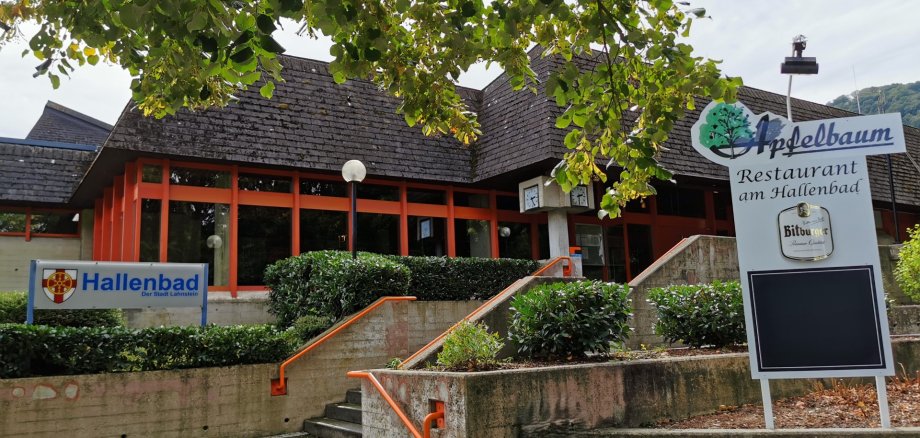 Hallenbad Lahnstein Außenansicht eines flachen Gebäudes mit dunklem Schrägdach und einer Fassade mit großen Fensterflächen und orangefarbenen Rahmen. Eine breite Treppenanlage mit orangefarbenen Handläufen führt zum Eingang hinauf. Vor dem Gebäude stehen Sträucher und Bäume. Links befindet sich ein Schild mit der Aufschrift „Hallenbad der Stadt Lahnstein“, rechts eine große Hinweistafel des Restaurants „Apfelbaum – Restaurant am Hallenbad“.