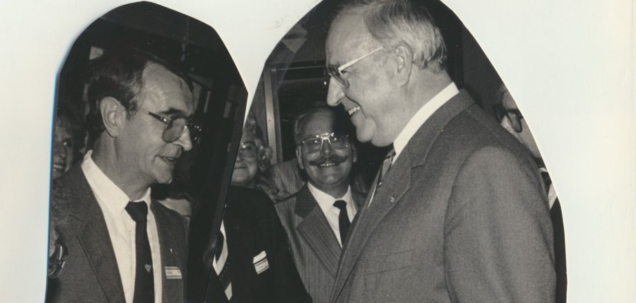Schwarz-Weiß-Foto: Zwei Männer in Anzügen, rechts steht der damalige Bundeskanzler Helmut Kohl, links der gewürdigte Hans Hoffmann aus Lahnstein, stehen einander bei einer Veranstaltung gegenüber und unterhalten sich. Im Hintergrund sind weitere lächelnde Personen zu sehen.