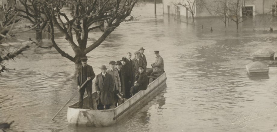 Mehrere Menschen stehen und sitzen in einem Boot, das durch ein überflutetes Gebiet fährt. Bäume und Häuser sind teilweise vom Hochwasser umgeben.