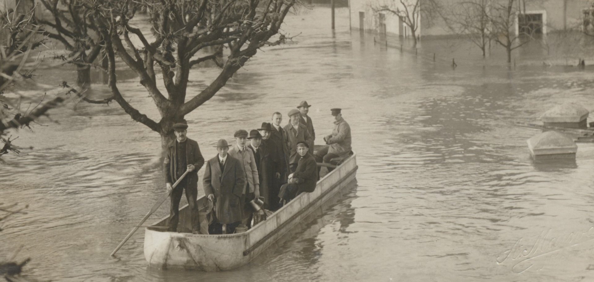 Mehrere Menschen stehen und sitzen in einem Boot, das durch ein überflutetes Gebiet fährt. Bäume und Häuser sind teilweise vom Hochwasser umgeben.